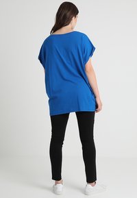 Žena s dlouhými hnědými vlasy, oblečená v volném modrém tričku s krátkým rukávem, černých úzkých kalhotách a bílých teniskách, stojící před jednoduchým světlým pozadím.