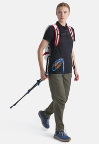 Attrezzatura da escursionismo: Una persona indossa un polo nera, pantaloni oliva e scarpe blu, porta un bastone da trekking blu e ha uno zaino leggero con spallacci rossi.