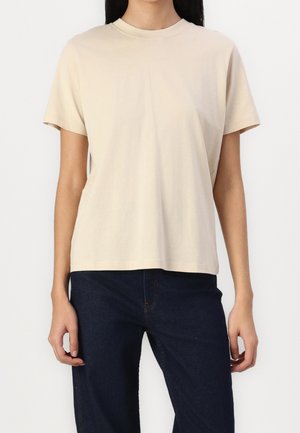 Femme portant un T-shirt beige uni à manches courtes, sorti par-dessus un jean bleu foncé, debout devant un fond blanc.