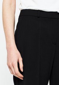 Zwarte op maat gemaakte broek van een gestructureerde stof, met een slanke tailleband en zijzakken. Hand op de heup geplaatst voor context.
