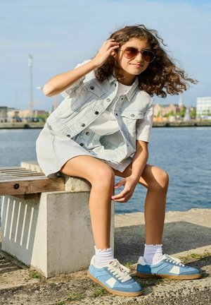 Gilet en denim bleu clair sur une chemise blanche, associé à une jupe gris clair. Baskets bleues avec des accents blancs et chaussettes à côtes.