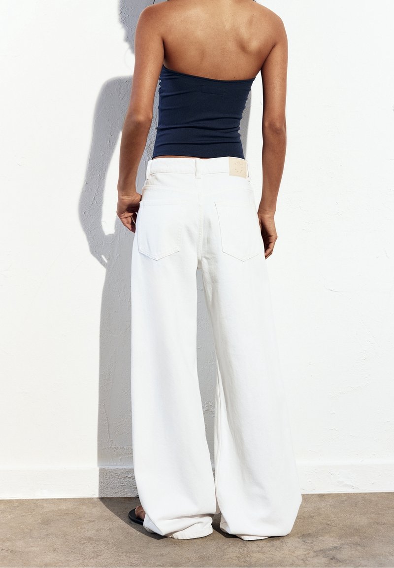 Personne mince portant un haut bustier bleu foncé et un pantalon blanc large et ample, debout contre un mur blanc.