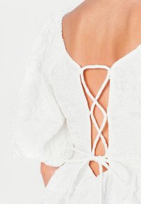 Abito bianco texturizzato con maniche a sbuffo, caratterizzato da un design incrociato sul retro e chiusura con lacci, che mette in risalto dettagli tessili intricati.