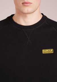 Black sweatshirt with a crew neckline, featuring a yellow "Barbour International" logo on the left chest. Fabric appears smooth and textured.
