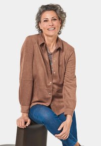 Giacca button-up in suede marrone con colletto, caratterizzata da una texture morbida e maniche lunghe, abbinata a jeans blu.