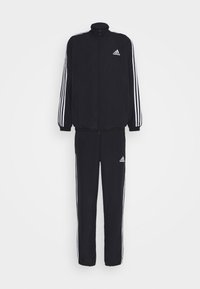 Survêtement Adidas noir avec trois bandes blanches sur les manches et les jambes, veste zippée et logos Adidas sur la poitrine et la cuisse.