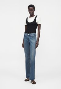 Mujer alta que lleva una camiseta negra de capas sobre una camiseta blanca de manga corta, vaqueros azules de pierna recta y sandalias negras con detalle de cadena dorada.