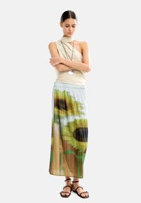 Jasno-beżowa bluzka zestawiona z plisowaną spódnicą w intensywny nadruk słoneczników. Modelka stoi z założonymi na siebie rękami, nosząc czarne sandały na paski.