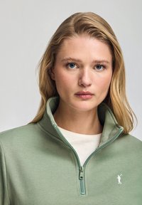 Grön zip-tröja med hög krage, med en vit logotyp på vänster sida och ett kontrasterande krämfärgat innerskikt.