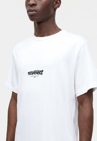Άνδρας που φοράει λευκό T-shirt The North Face με μαύρο λογότυπο στο κέντρο του στήθους, σε απλό φόντο.