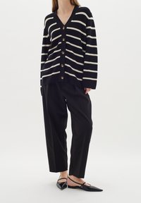 Žena v černobíle pruhovaném zapínacím kardiganu, volných černých kalhotách a černých špičatých botách s otevřenou patou na bílém pozadí.