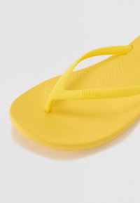 Keltainen flip-flop sandaali, jossa on teksturoitu pohja, sileät hihnat ja logo sivulla. Valmistettu joustavasta materiaalista ja minimalistisesta muotoilusta.