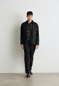 Jachetă din denim negru cu închidere cu butoane și două buzunare frontale, asociată cu pantaloni negri asortați și o cămașă cu dungi. Textură netedă.