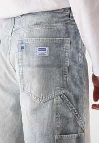 Jeans din denim cu dungi, de culoare albastru deschis, cu un buzunar posterior. Prezintă o etichetă din material pe care scrie "HUGO" și detalii de cusătură contrastante.