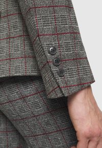 Grijs geruite blazer met een mouw die een gestructureerde stof toont met een rood en zwart ruitpatroon, afgewerkt met vier zwarte knopen bij de manchet.
