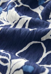 Marinfärgad tyg med en texturerad yta, som har abstrakta vita och ljusblå blommönster. Materialet verkar lätt och något skrynkligt.