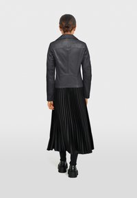 Čierna kožená bunda s priliehavým strihom, zúženým golierom a viditeľným šitím, skombinovaná s dlhou čiernou plisovanou sukňou a čiernymi čižmami.