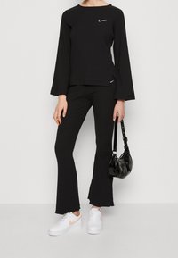 Person som har på sig en svart ribbad långärmad topp och vida byxor, vita sneakers med rosa swoosh, och håller en liten svart handväska.