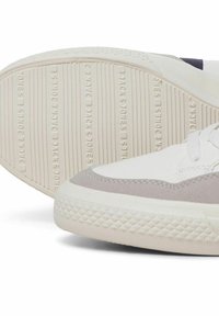 Witte sneaker met grijze suède accenten, textuur rubberen zool met verticale strepen en branding, en ronde veters op een gladde bovenzijde.