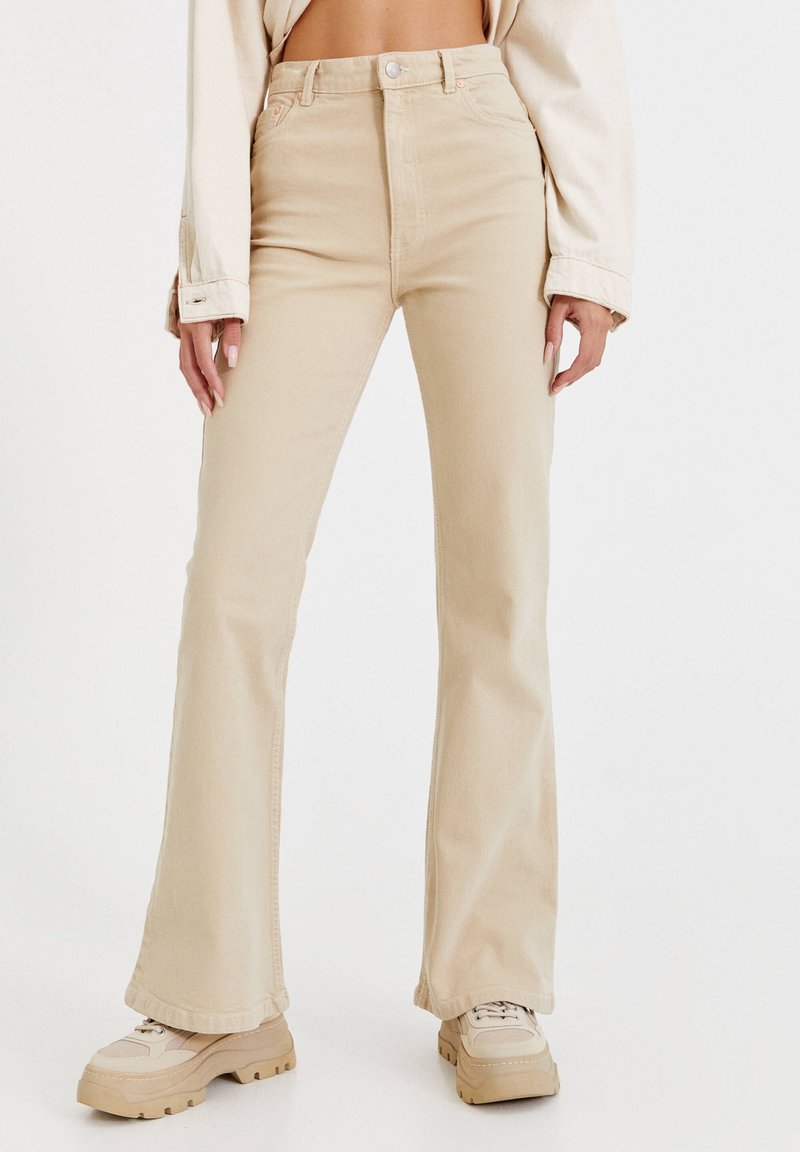 Femme portant un pantalon évasé taille haute beige, des chaussures à plateforme épaisses assorties et un top court beige à manches longues.