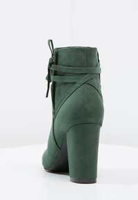 Botín de ante verde con tacón alto, que presenta un diseño de cordones y correas cruzadas en la parte trasera, textura suave y puntera redondeada.