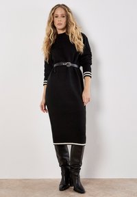 Schwarzes Strickkleid mit einem gerippten Saum und weißen Streifen an den Ärmeln, tailliert mit einem schwarzen Ledergürtel. Schwarze kniehohe Stiefel.