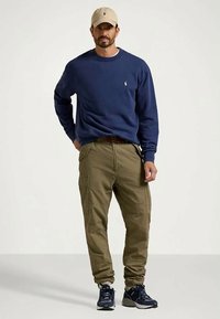 Άνδρας που φορά καπέλο μπεζ, σκούρα μπλε φούτερ, ελαιόλαδο cargo παντελόνια, καφέ ζώνη και σκούρα μπλε αθλητικά παπούτσια, στέκεται με το ένα χέρι στην τσέπη.