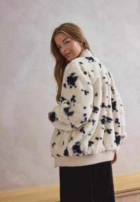 Cream faux fur bomber jacket with black spots, ribbed cuffs, and hem. Soft texture, slightly oversized fit. Model poses from the back.