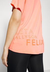 Camisola desportiva cor-de-corais com texto em laranja bold a dizer "LETA ALLYSON FELIX." As mangas curtas e o corte descontraído são visíveis. Combinada com leggings pretas.