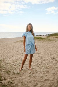 Femme portant un haut bleu clair à manches courtes à volants et un short assorti, debout sur une plage de sable avec la mer et un ciel nuageux en arrière-plan.