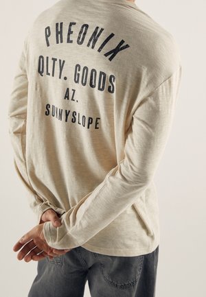 Chemise beige à manches longues avec un texte noir « Pheonix Qlty. Goods AZ Sunnyslope » portée par une personne avec les mains jointes dans le dos.