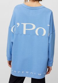 Överdimensionerad blå sweatshirt i bomull, med vit grafisk text: "c'Pol" på baksidan och "STOCKHOLM '21" vid fållen.