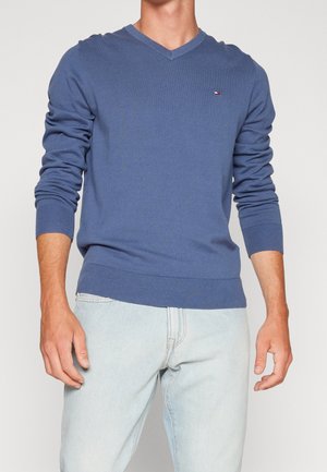 Homme portant un pull bleu à col en V à manches longues et un jean bleu clair, sur un fond uni.