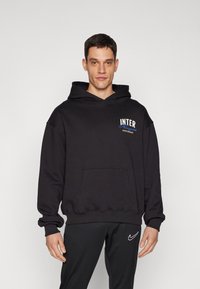 Hoodie preto com um bolso canguru, apresentando um logo "INTER" em branco e azul. Tecido texturizado, corte solto, design com capuz em camadas. Calças pretas.