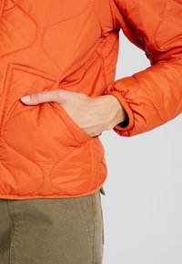 Main dans la poche d'une veste matelassée orange vif avec des coutures en forme de vague, associée à un pantalon vert olive.