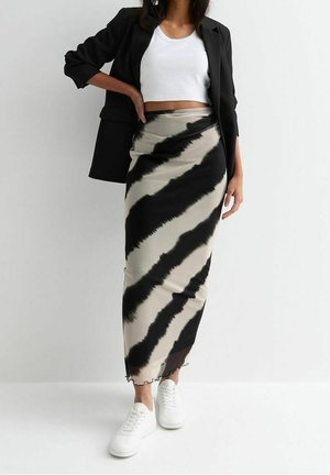 Femme portant un blazer noir, un top court blanc, une longue jupe à rayures diagonales noires et beige, et des baskets blanches, debout contre un mur blanc.