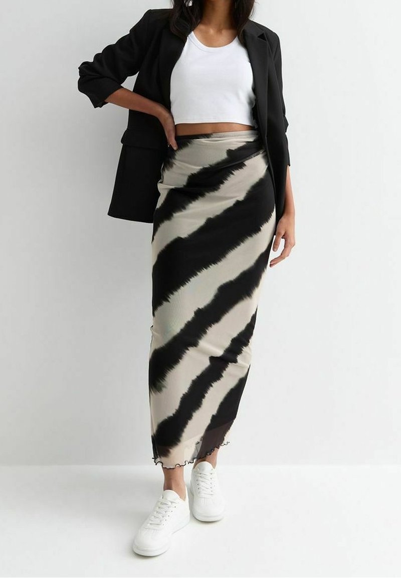Femme portant un blazer noir, un top court blanc, une longue jupe à rayures diagonales noires et beige, et des baskets blanches, debout contre un mur blanc.