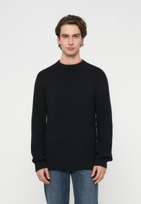 Pulover negru din tricot cu mâneci raglan, guler rotund, manșete și tiv dantelat. Purtați cu blugi albaștri pe un fundal alb simplu.