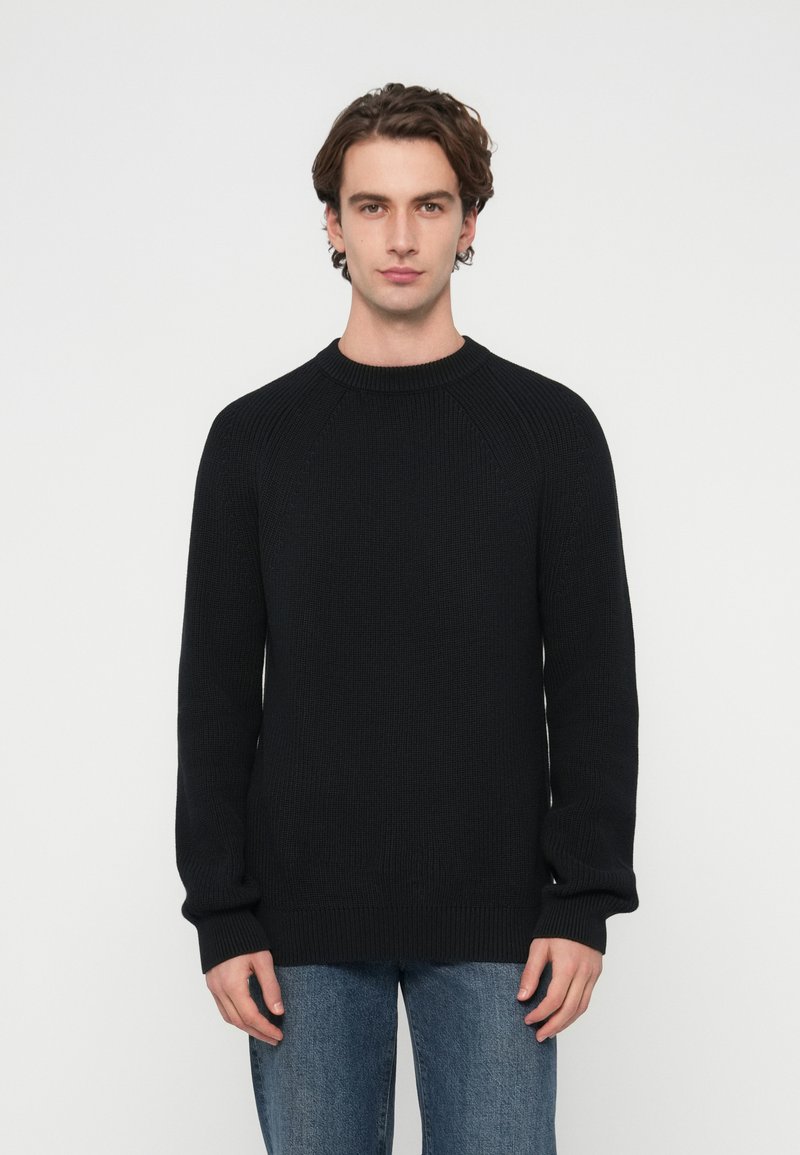 Pulover negru din tricot cu mâneci raglan, guler rotund, manșete și tiv dantelat. Purtați cu blugi albaștri pe un fundal alb simplu.