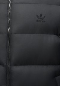 Zwarte doorgestikte jas met centrale ritssluiting en zwart Adidas-logo op de linkerborst.