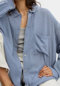 Chemise bleu clair boutonnée en tissu doux et texturé, avec une seule poche poitrine et une coupe décontractée. Manches longues avec poignets boutonnés.