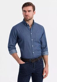 Donkerblauwe button-up shirt met wit stippenpatroon, opgerolde mouwen en een borstzak, gecombineerd met donkere jeans en een bruine riem.