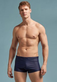 Marineblaue Badehose aus glattem Material, mit taillierter Passform und elastischem Bund, auf einem oberkörperfreien Modell vor einem hellen Hintergrund präsentiert.