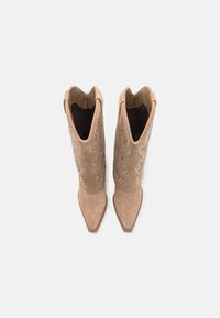 Botas de vaquero de ante color marrón claro con puntas afiladas, que cuentan con costuras decorativas y adornos a lo largo del caño. Forro de tela marrón en el interior.