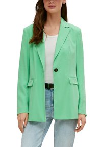 Minzgrüner Blazer mit einem einzelnen braunen Knopf, strukturierten Revers und Klappentaschen; getragen über einem weißen Oberteil und hellblauen Jeans.