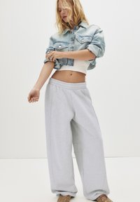 Frau in einer kurz geschnittenen hellen Jeansjacke, einem weißen Tanktop, lockeren hellgrauen Jogginghosen und beigen Sandalen mit Lochdetails.