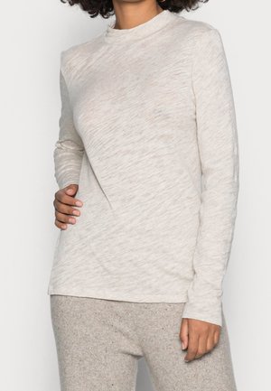 Woman wearing light beige long-sleeve top and matching knit pants, standing with one hand on hip and the other relaxed by side.