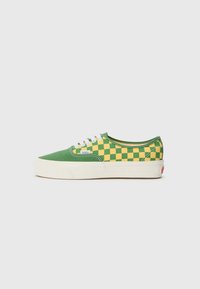 Green canvas sneakers featuring a yellow checkerboard pattern, white laces, and a cream rubber sole with a red logo tab on the heel.