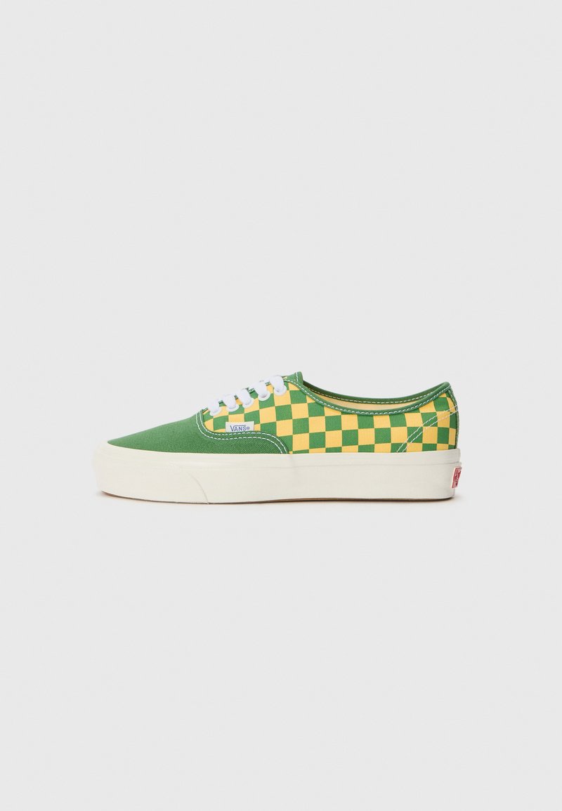 Green canvas sneakers featuring a yellow checkerboard pattern, white laces, and a cream rubber sole with a red logo tab on the heel.