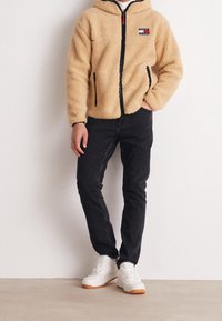 Persona che indossa una giacca in pile beige con zip e logo Tommy Jeans, jeans neri e sneakers bianche, in piedi con una mano nella tasca.
